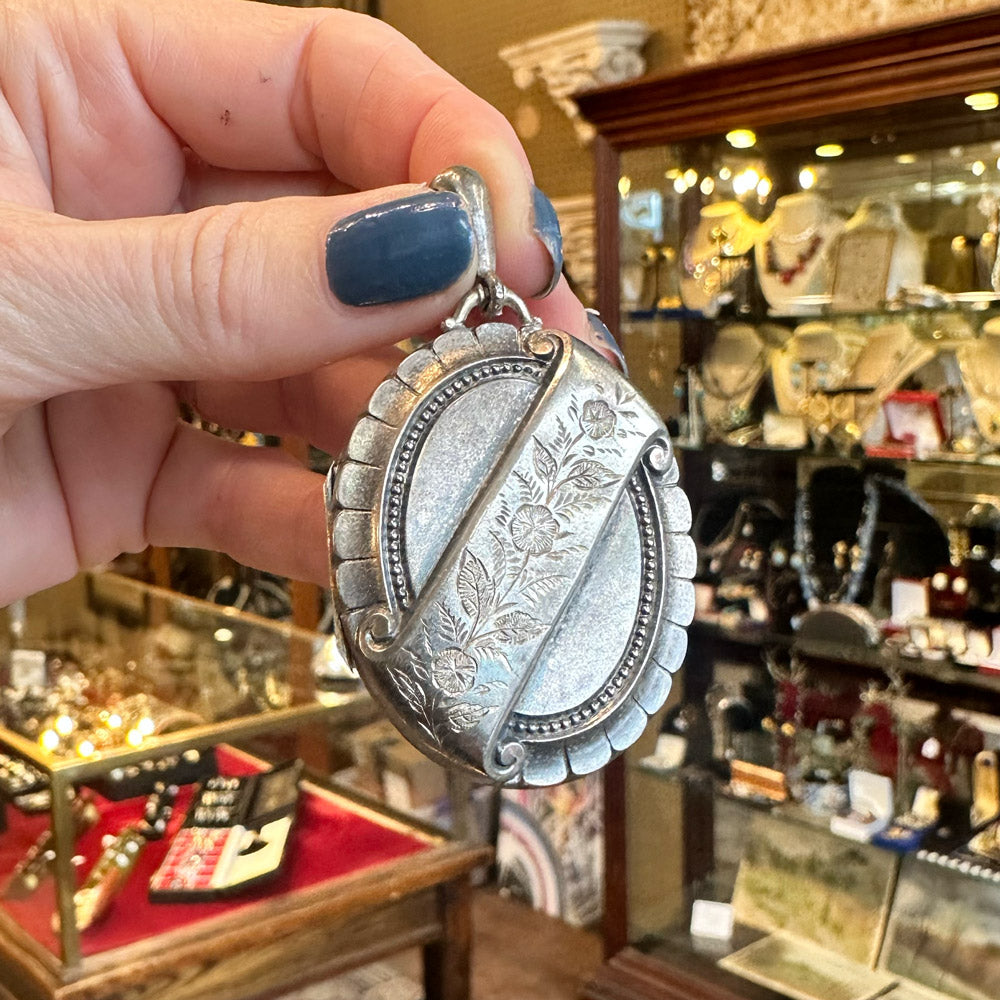 Silver locket with engraving held by a hand with blue nail polish inside an antique jewelry store.