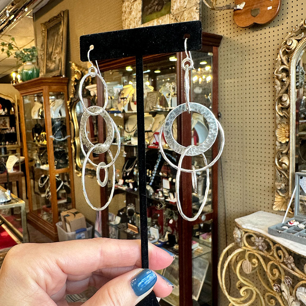 Hammered, textured, silver hoop earrings held by a hand in front of a jewelry store display.