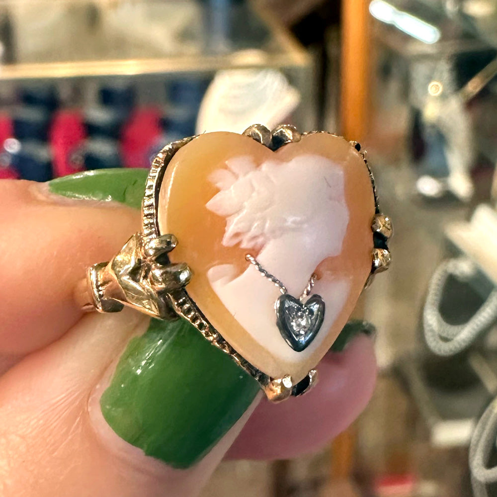 Heart-shaped cameo ring with gold frame held by a hand with green nail polish inside an antiques store.