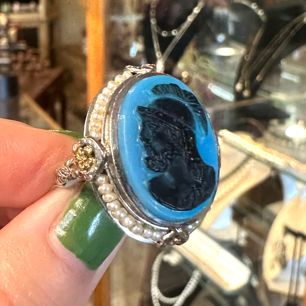 Art Deco Agate and Pearl ring with blue stone and black engraving held by a hand with green nail polish, blurred antique store background.
