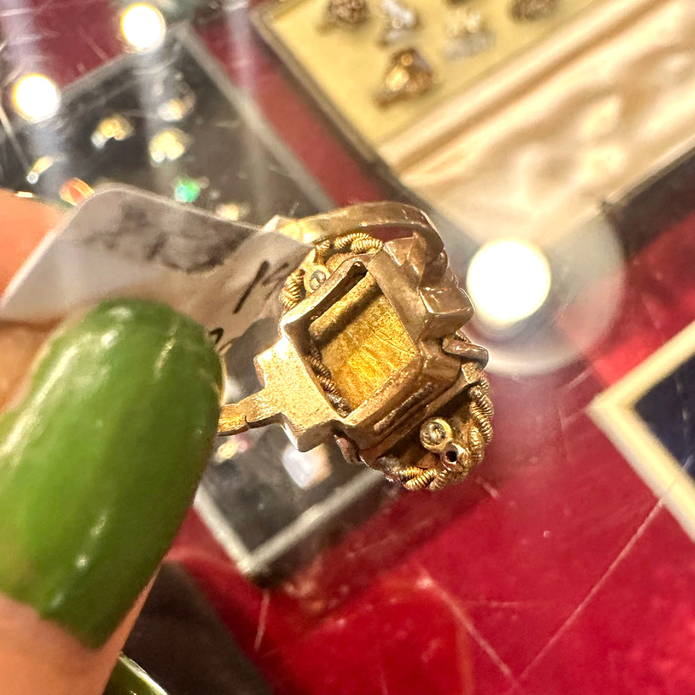 Close-up of the back of a gold ring with intricate design held by a hand against a blurred background.