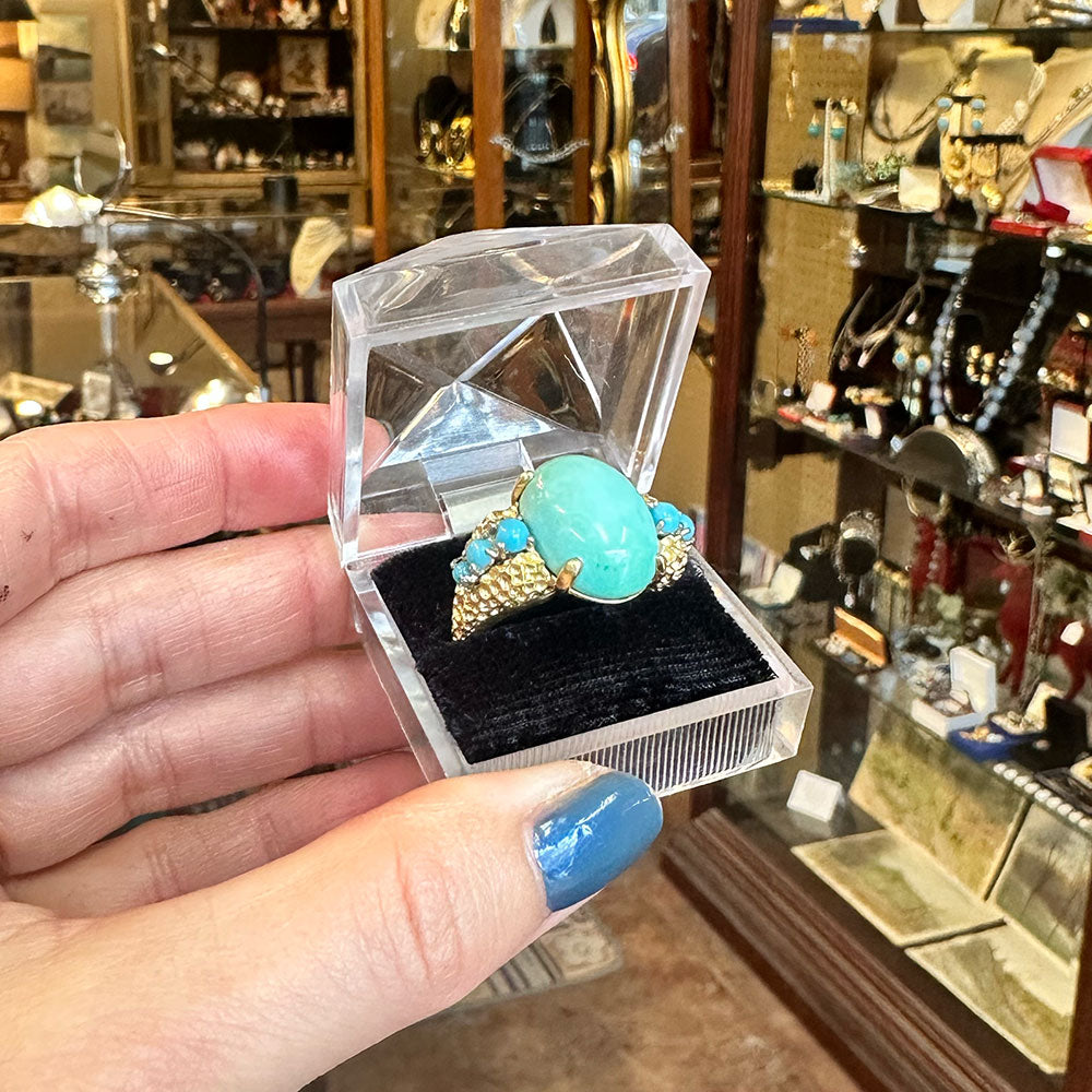 Mid-Century vintage turquoise ring in a clear box held by a hand with blue nail polish, in a store setting.
