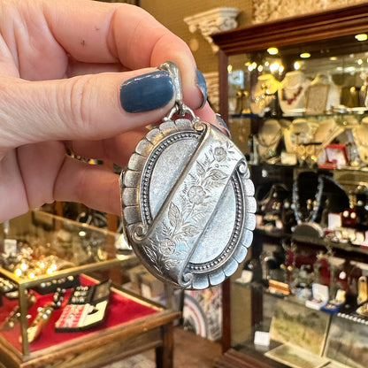Silver locket with engraving held by a hand with blue nail polish inside an antique jewelry store.