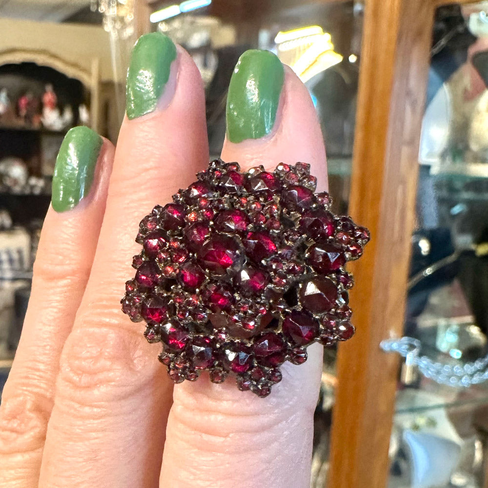 Close-up of a hand wearing a large, Victorian ring with red gemstones and gold against a blurred background.