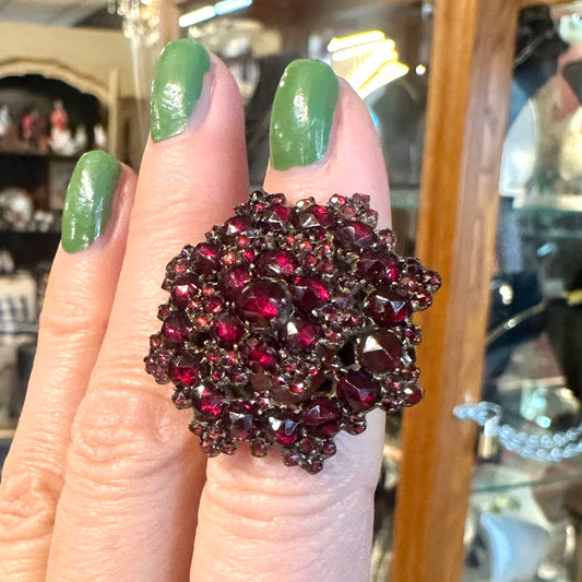 Close-up of a hand wearing a large, Victorian ring with red gemstones and gold against a blurred background.