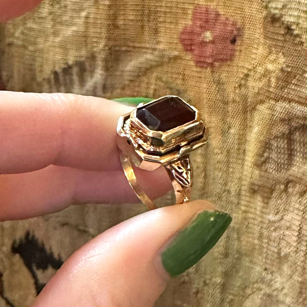 Gold flip ring from the 20s with a red gemstone held up against a floral fabric background inside an antique store.