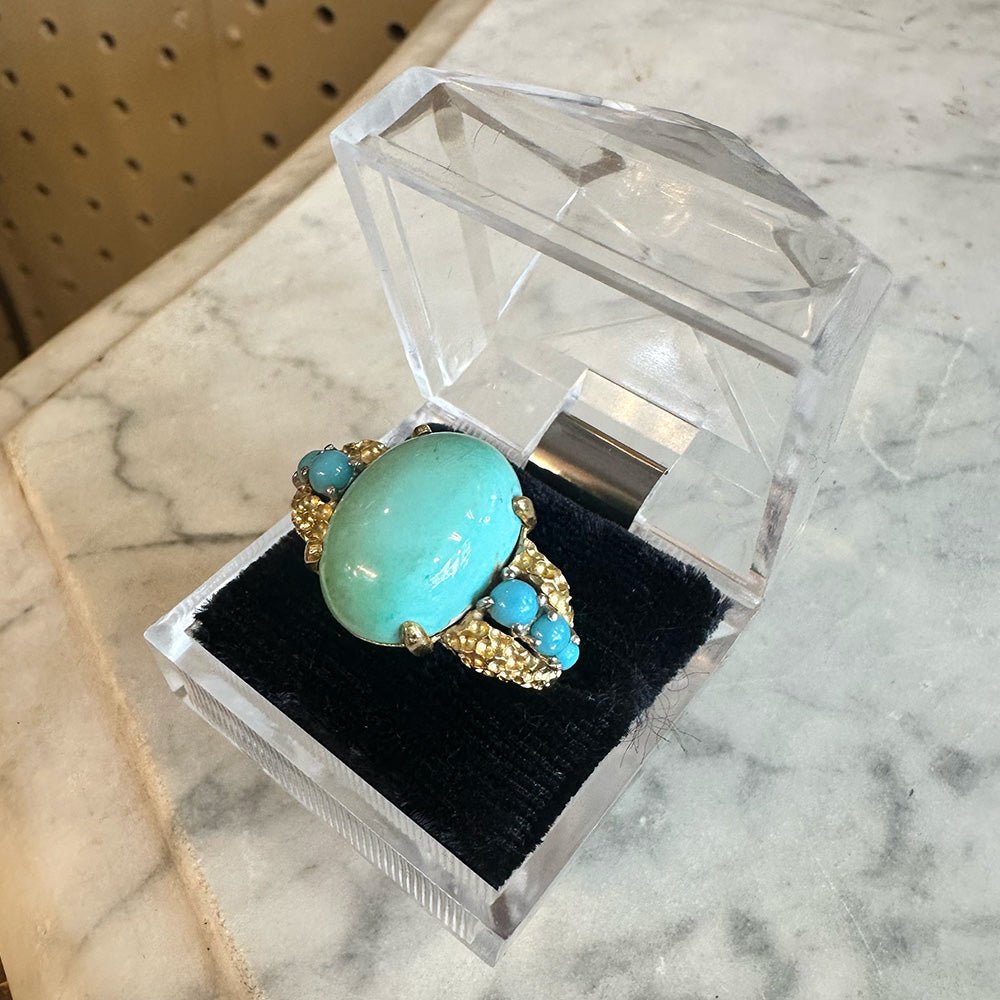 Gold ring with turquoise stones on a black velvet cushion in a clear jewelry box sitting on a marble surface.
